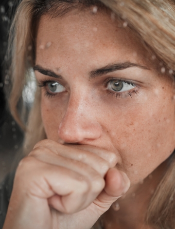Blonde woman anxiously looks out a rainy window with her fist covering her mouth