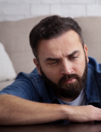 Bearded man sitting on the ground at a wooden table with a couch behind him. He looks deep in thought and his arm rests on the table in front of him