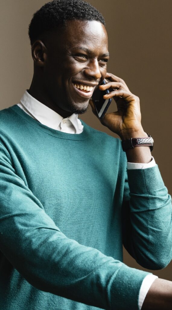 Dark skinned man wearing a green sweater and white button-up shirt underneath smiling and talking on the phone